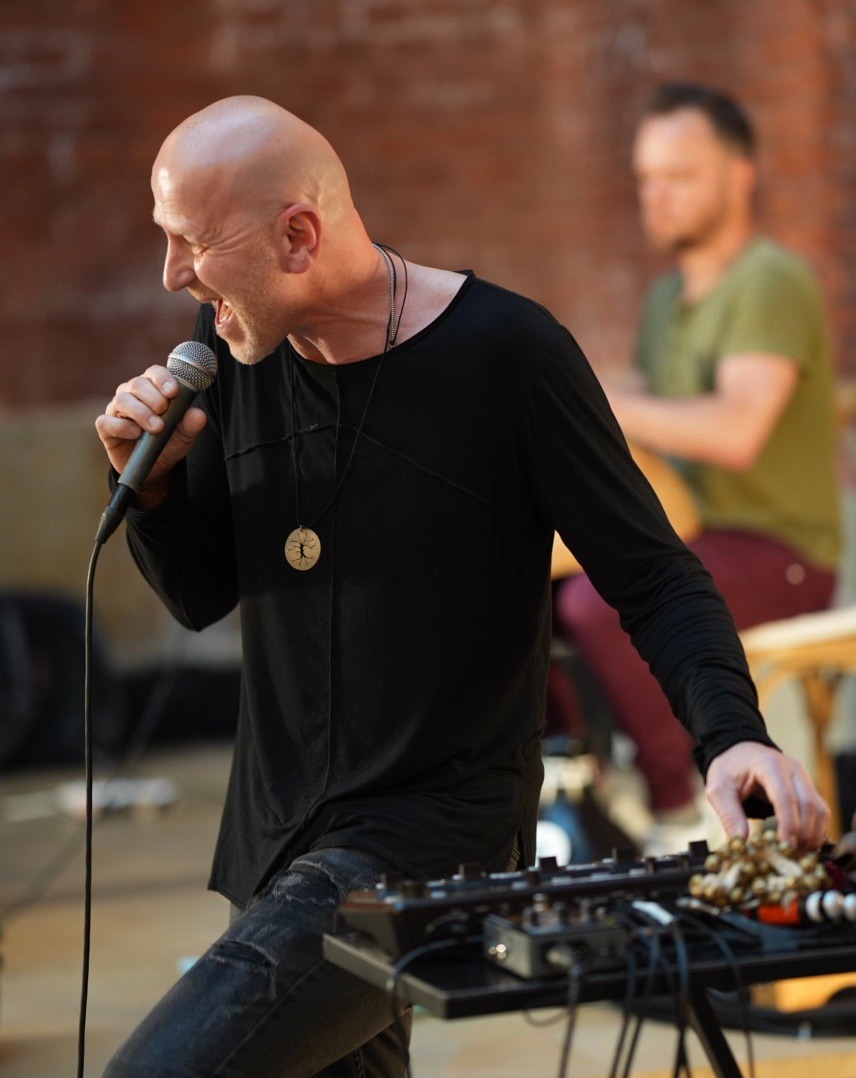 Wokalista w czarnej koszuli z długim rękawem śpiewa do mikrofonu, obok syntezator i drugi muzyk w tle.