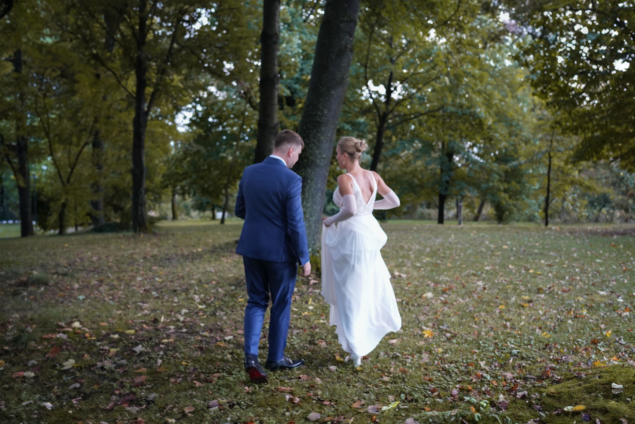 Para młoda spaceruje po parku w jesiennej scenerii, panna młoda w sukni z odkrytymi plecami i długimi rękawiczkami, pan młody w granatowym garniturze, widok z tyłu.
