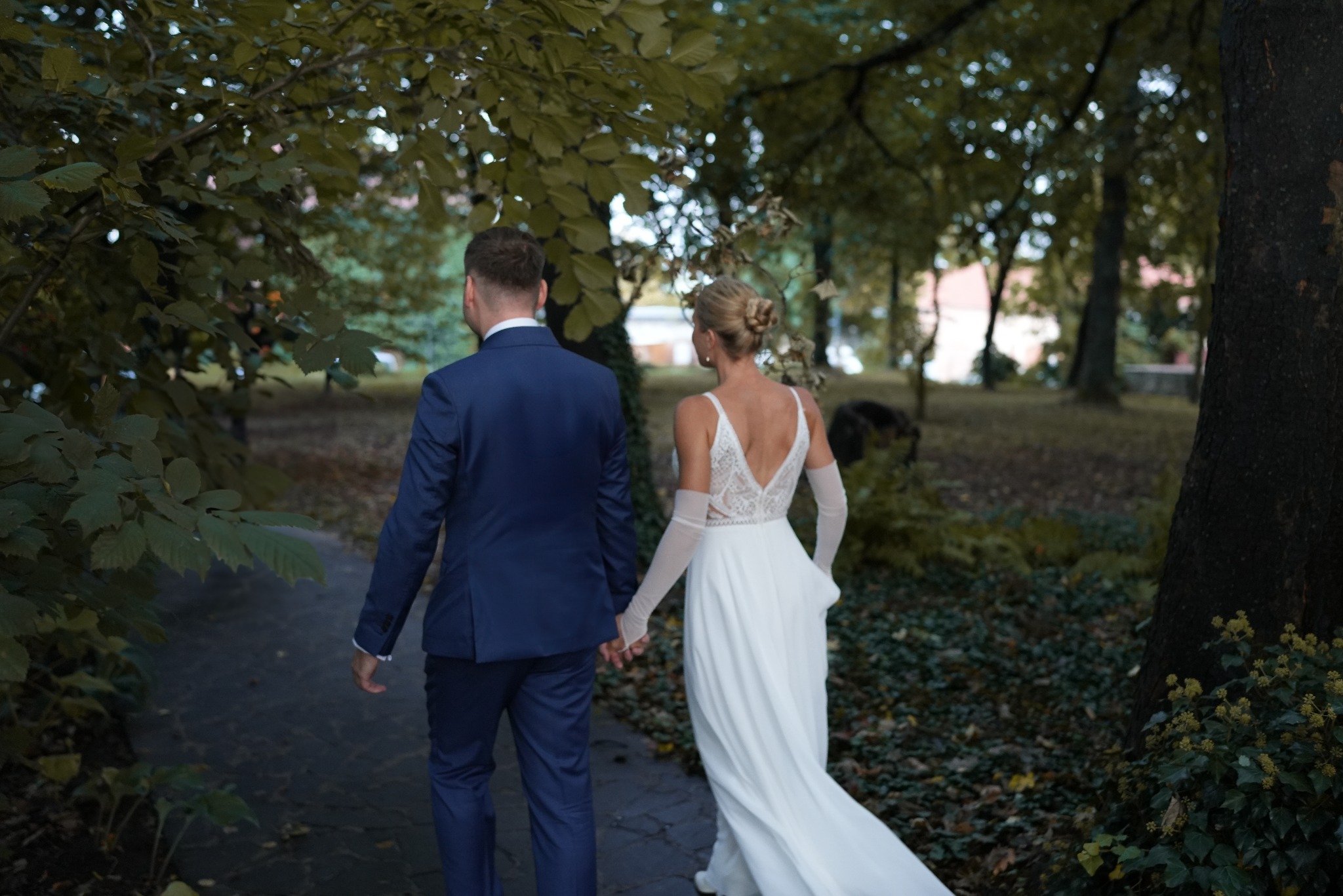 Para młoda trzymająca się za ręce, spacerująca parkową alejką, panna młoda w sukni z koronkowym wykończeniem pleców i długich, przezroczystych rękawiczkach, pan młody w granatowym garniturze.