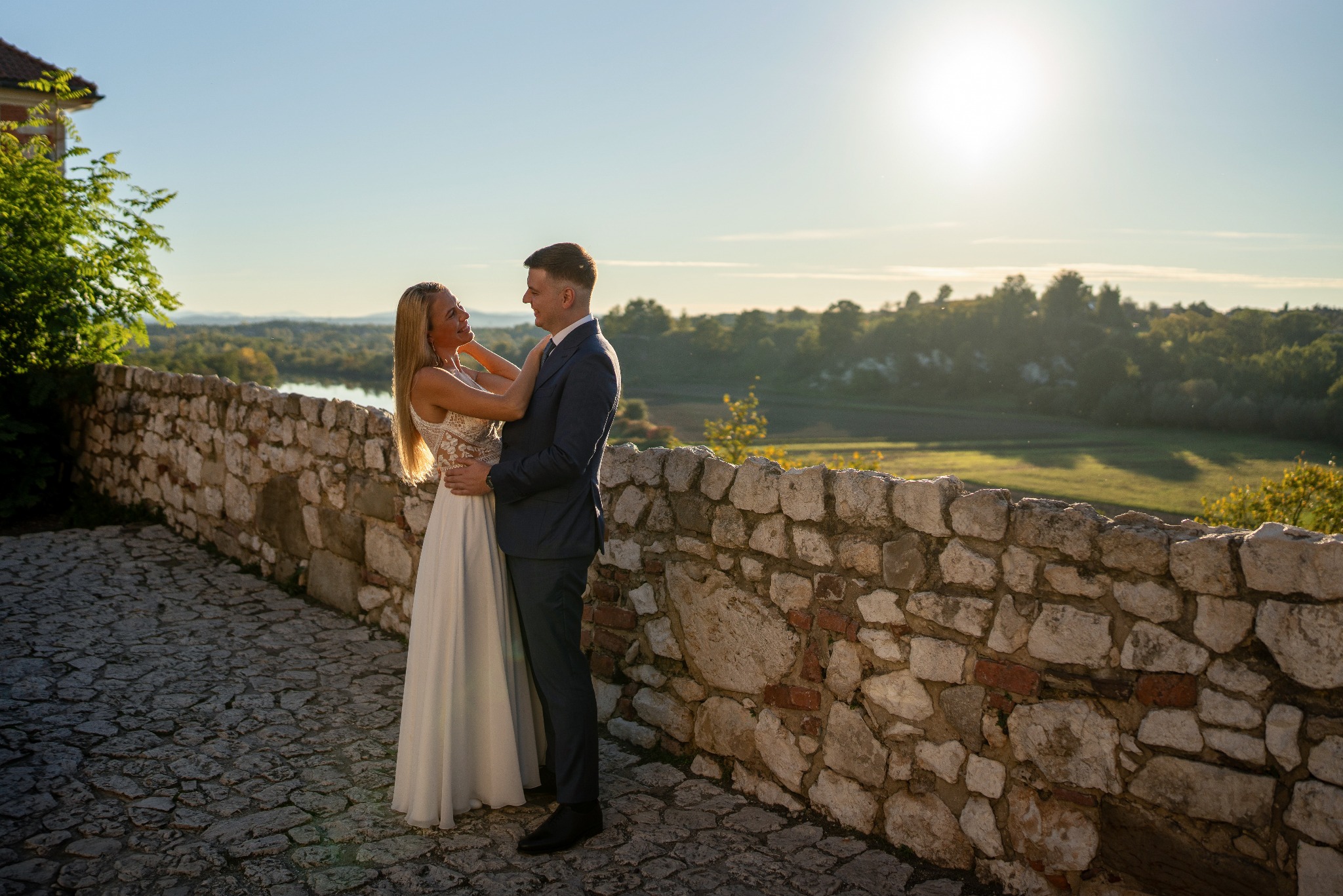 Para młoda w objęciach na tle kamiennego muru i rozległego krajobrazu w ciepłym świetle zachodzącego słońca.