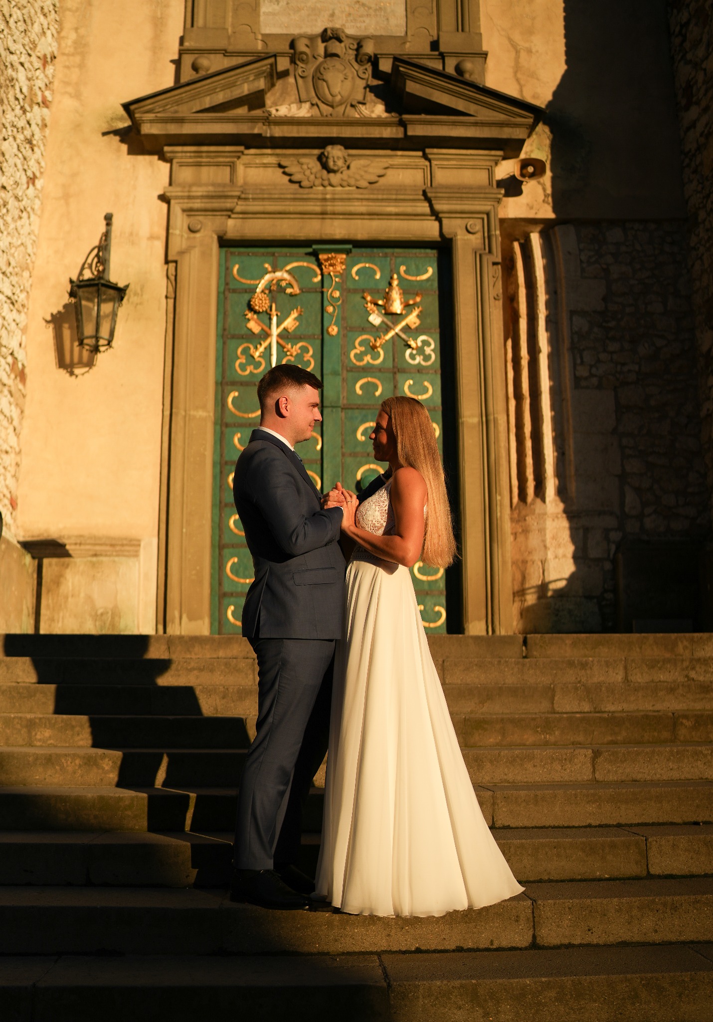 Para młoda trzymająca się za ręce na schodach przed ozdobnymi, zielonymi drzwiami w stylu vintage, ciepłe, zachodzące słońce oświetla budynek.