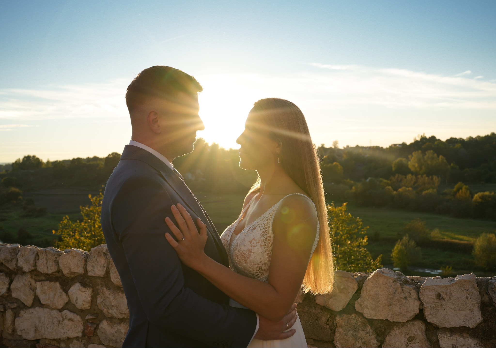 Para młoda w objęciach na tle zachodzącego słońca, sylwetki na tle zielonych wzgórz, kamienny murek w dolnej części kadru.