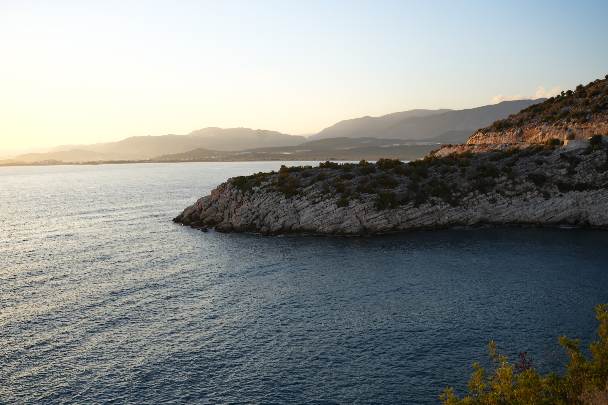 Spokojne morze obmywa skalisty klif porośnięty zielenią, w oddali zarys gór w miękkim, zachodzącym słońcu.