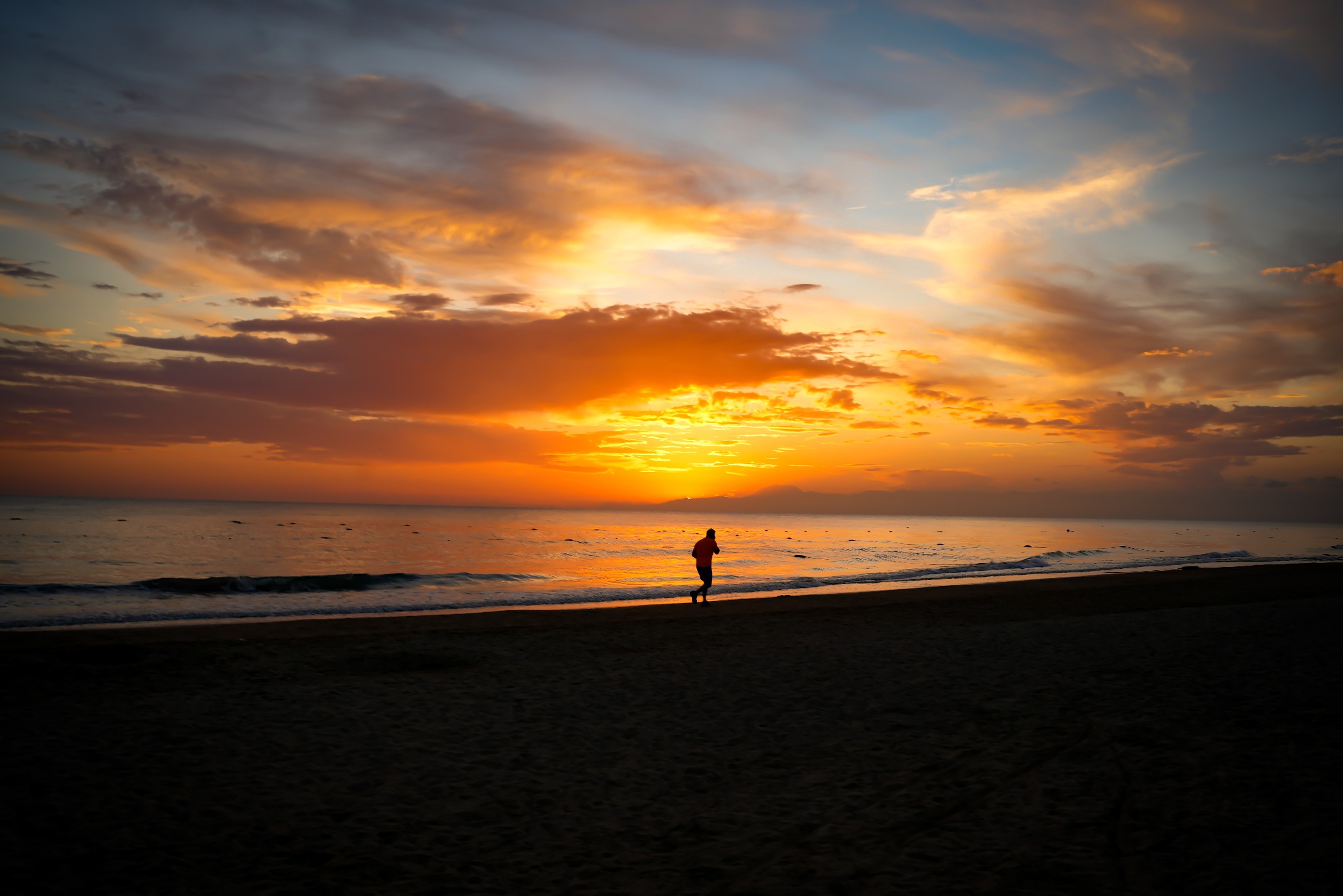 Sylwetka biegacza na plaży o wschodzie słońca, morze odbija pomarańczowe niebo z chmurami.