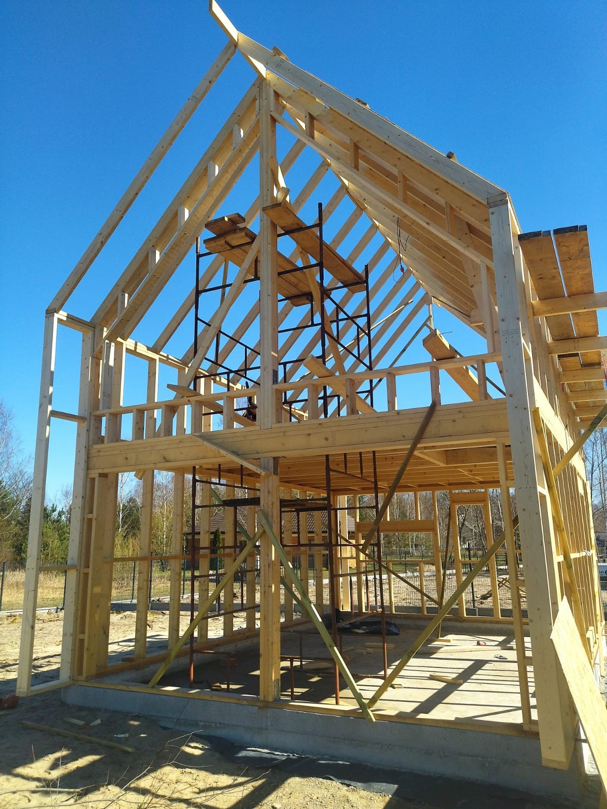 Konstrukcja drewniana domu w trakcie budowy, widoczne rusztowania i fundamenty, błękitne niebo w tle.