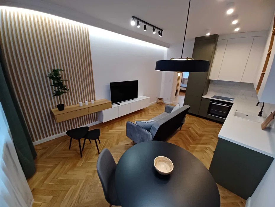 Nowoczesne wnętrze mieszkania: parkiet w jodełkę, ściana z drewnianych listew, telewizor na białej szafce, szara sofa, okrągły czarny stół, zielono-biała zabudowa kuchenna z marmurowym blatem.