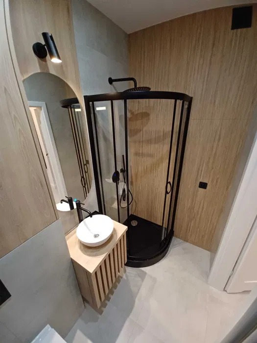 Nowoczesna łazienka z kabiną prysznicową w czarnej ramie, okrągłą umywalką nablatową na drewnianej szafce z lamelami i lustrem z oświetleniem punktowym.