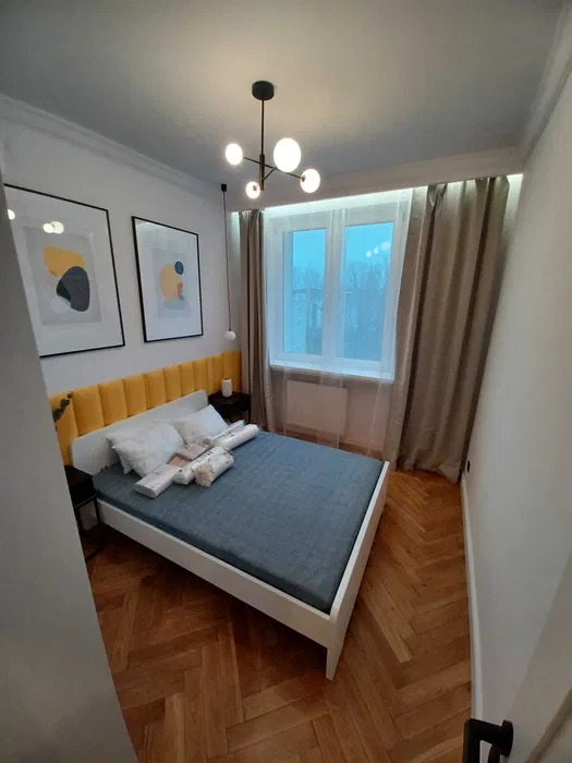 Nowoczesna sypialnia z parkietem w jodełkę, łóżkiem z żółtym zagłówkiem i minimalistyczną lampą wiszącą.