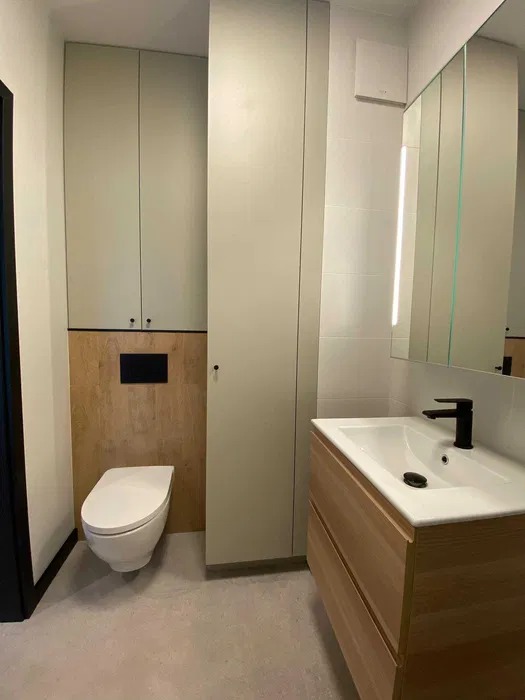 Nowoczesna łazienka w Warszawie z podwieszaną toaletą, drewnianą zabudową umywalki z czarną armaturą i wysoką szafką w neutralnym odcieniu.