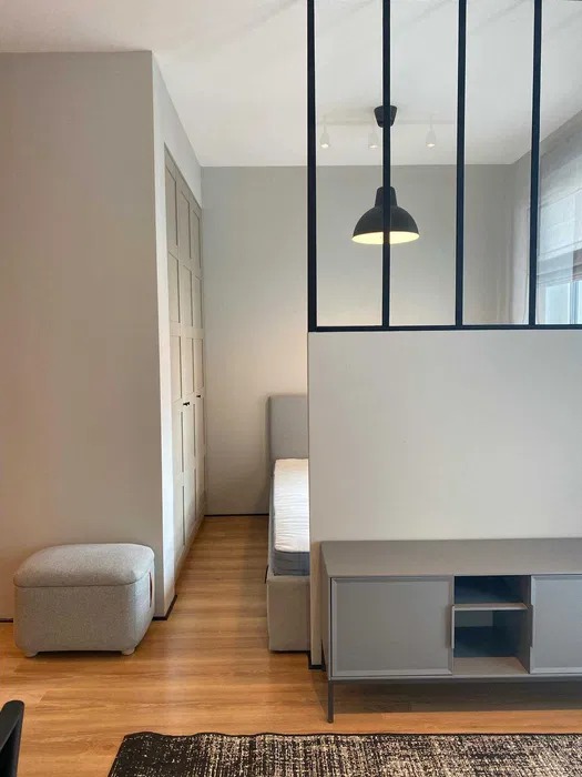 Nowoczesna sypialnia z drewnianą podłogą, szarą komodą i łóżkiem, oddzielona od reszty pomieszczenia ścianką działową z czarnymi, metalowymi profilami i wiszącą lampą.