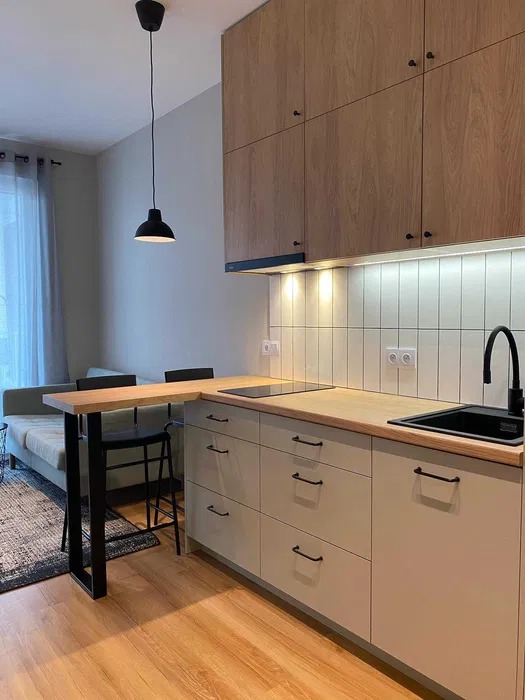 Nowoczesna kuchnia w jasnych barwach z drewnianymi akcentami, czarną armaturą i lampą wiszącą, widok na salon z sofą i dywanem.