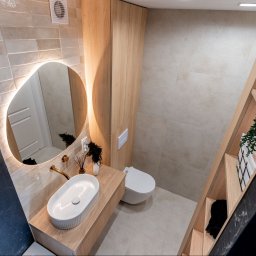 ARCHITEKTURA WNĘTRZ Jakub Tarnowski - Nowoczesna łazienka z owalnym lustrem LED, drewnianymi szafkami i umywalką nablatową. Beżowe płytki i czarny marmur tworzą elegancki kontrast.