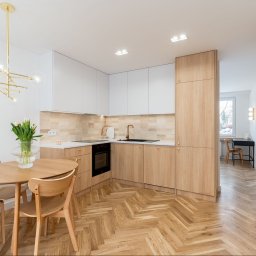 ARCHITEKTURA WNĘTRZ Jakub Tarnowski - Jasna kuchnia z drewnianą podłogą ułożoną w jodełkę, białe szafki górne i drewniane dolne. Na stole wazon z tulipanami i złoty żyrandol.