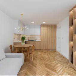 Jasne, nowoczesne wnętrze mieszkania z parkietem w jodełkę, okrągłym stołem, szarą kanapą i zabudową kuchenną z elementami drewna. Widok na regał z podświetleniem.