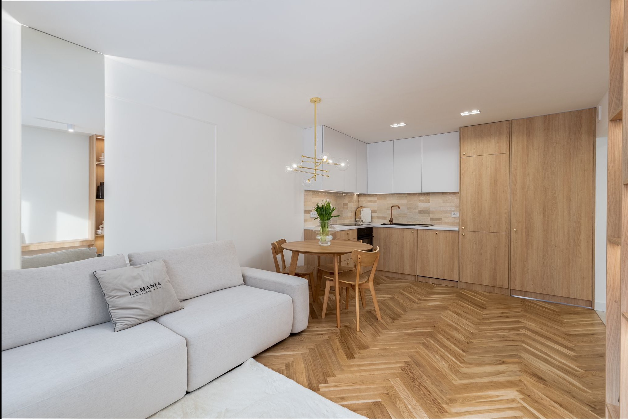 Jasne wnętrze: sofa z poduszką 'La Mania', okrągły stół z tulipanami, parkiet w jodełkę i zabudowa kuchenna z drewnianymi frontami. Minimalistyczny design.
