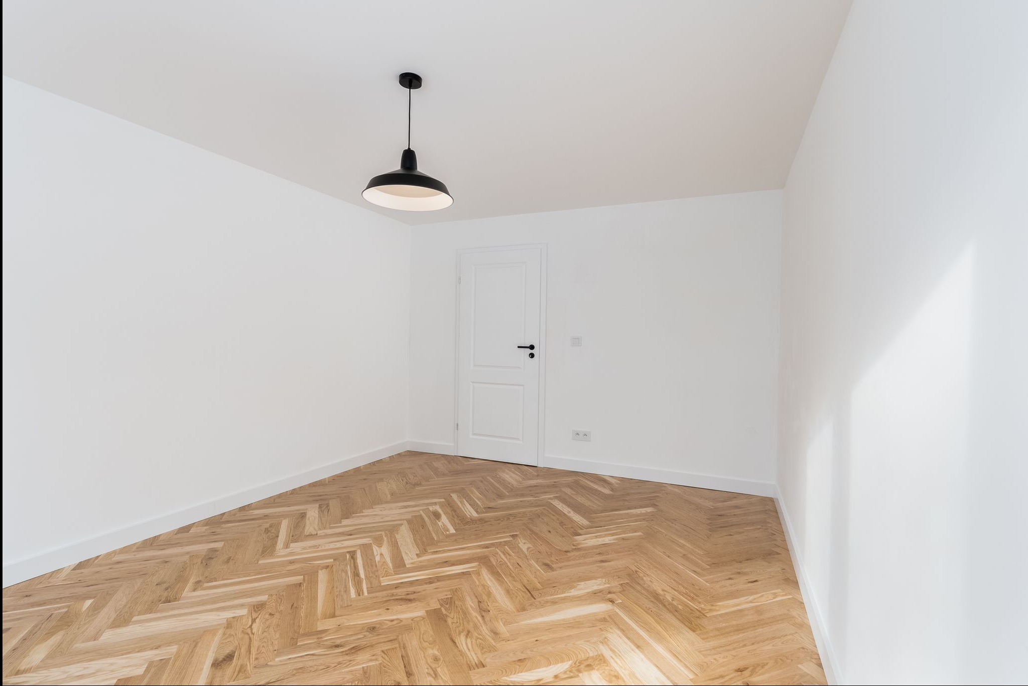 Pusty pokój z parkietem ułożonym we wzór jodełki, białe ściany, białe drzwi i czarna lampa wisząca. Minimalistyczne wnętrze po remoncie.