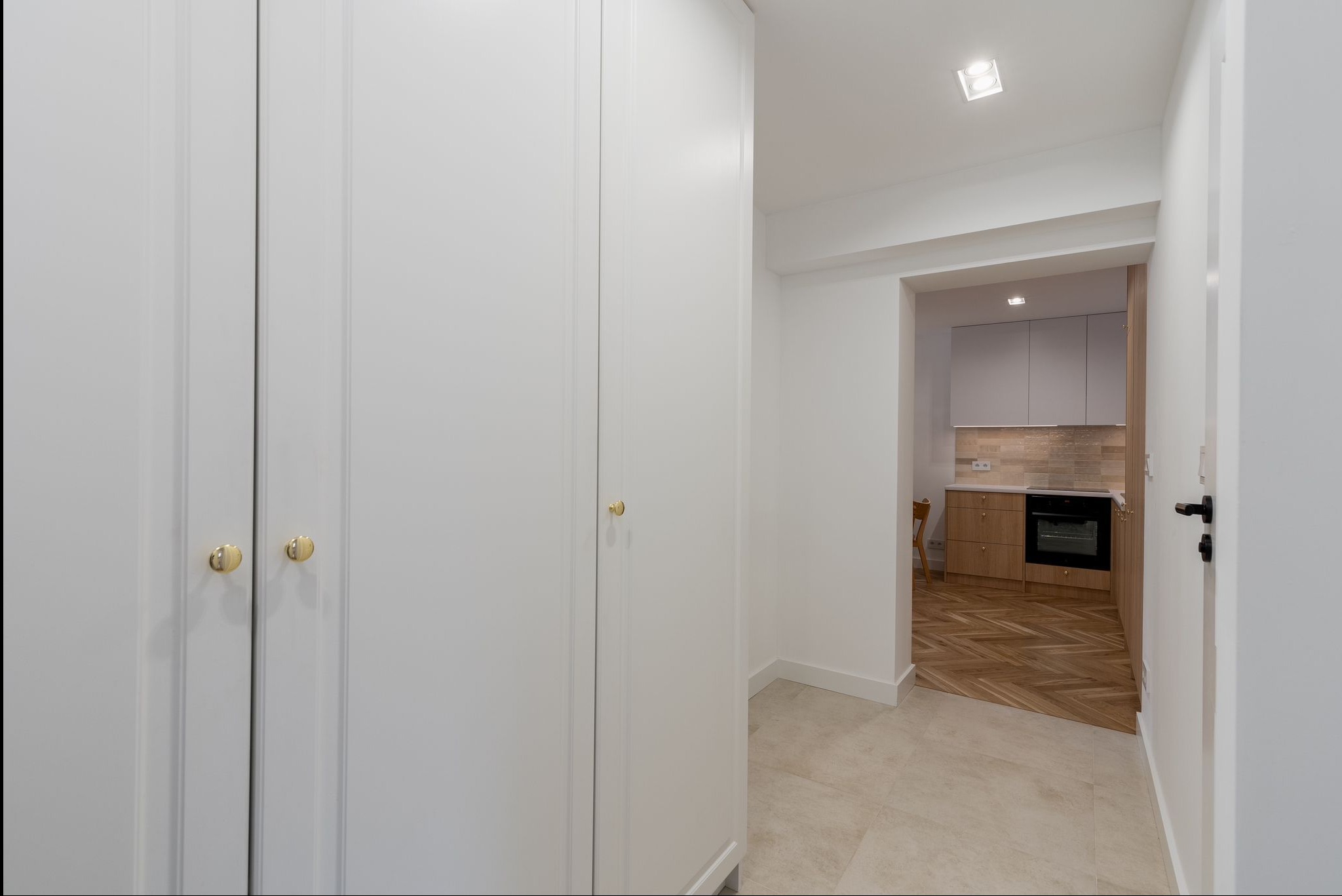 Wnętrze: biała szafa w zabudowie z złotymi uchwytami, fragment kuchni z drewnianą podłogą w jodełkę w tle, jasne płytki na podłodze w przedpokoju.