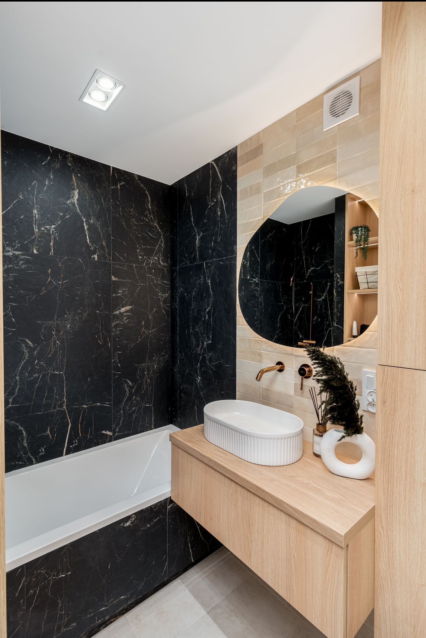 Nowoczesna łazienka z czarnymi płytkami imitującymi marmur, drewnianą szafką i owalnym lustrem z podświetleniem. Minimalistyczny design.
