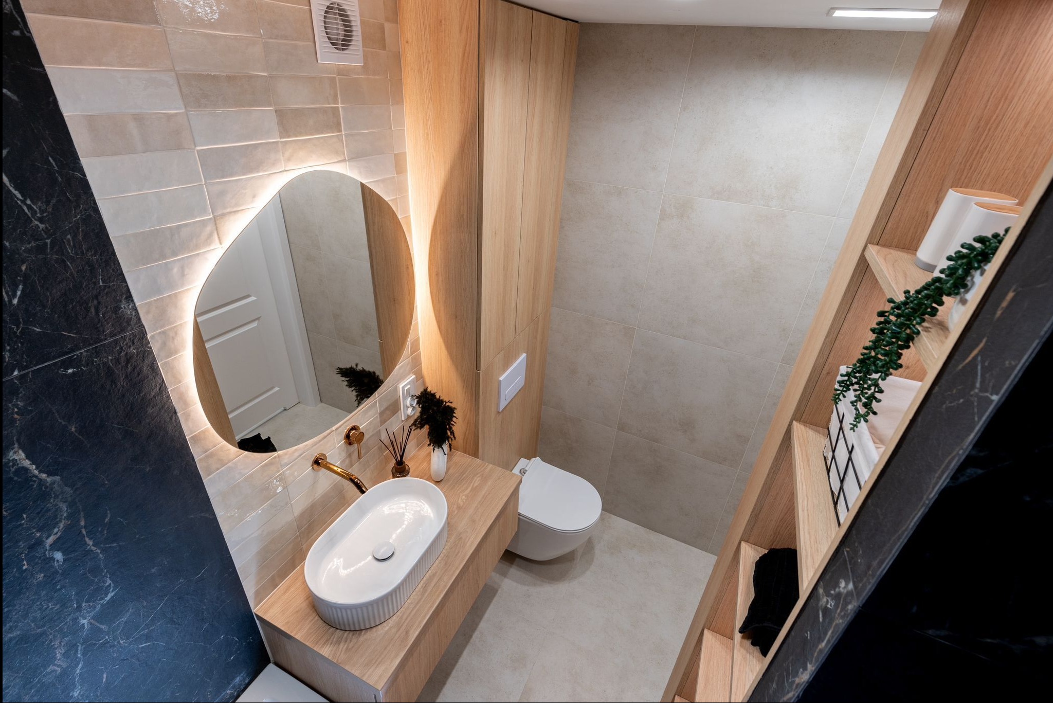 Nowoczesna łazienka z owalnym lustrem LED, drewnianymi szafkami i umywalką nablatową. Beżowe płytki i czarny marmur tworzą elegancki kontrast.