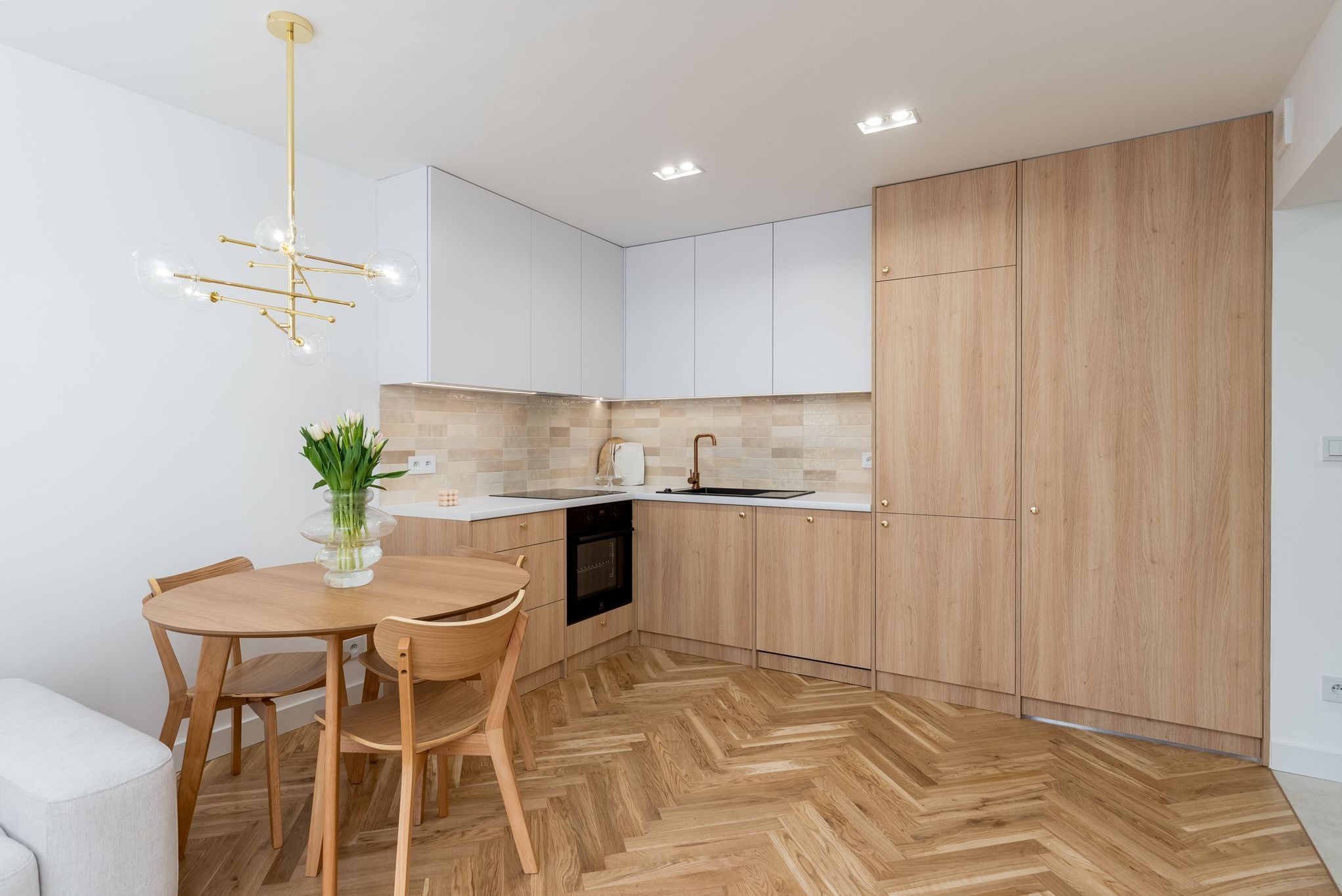 Jasna kuchnia z drewnianą podłogą w jodełkę, okrągłym stołem i złotymi akcentami: lampa, bateria. Białe fronty szafek górnych i drewniane dolne.