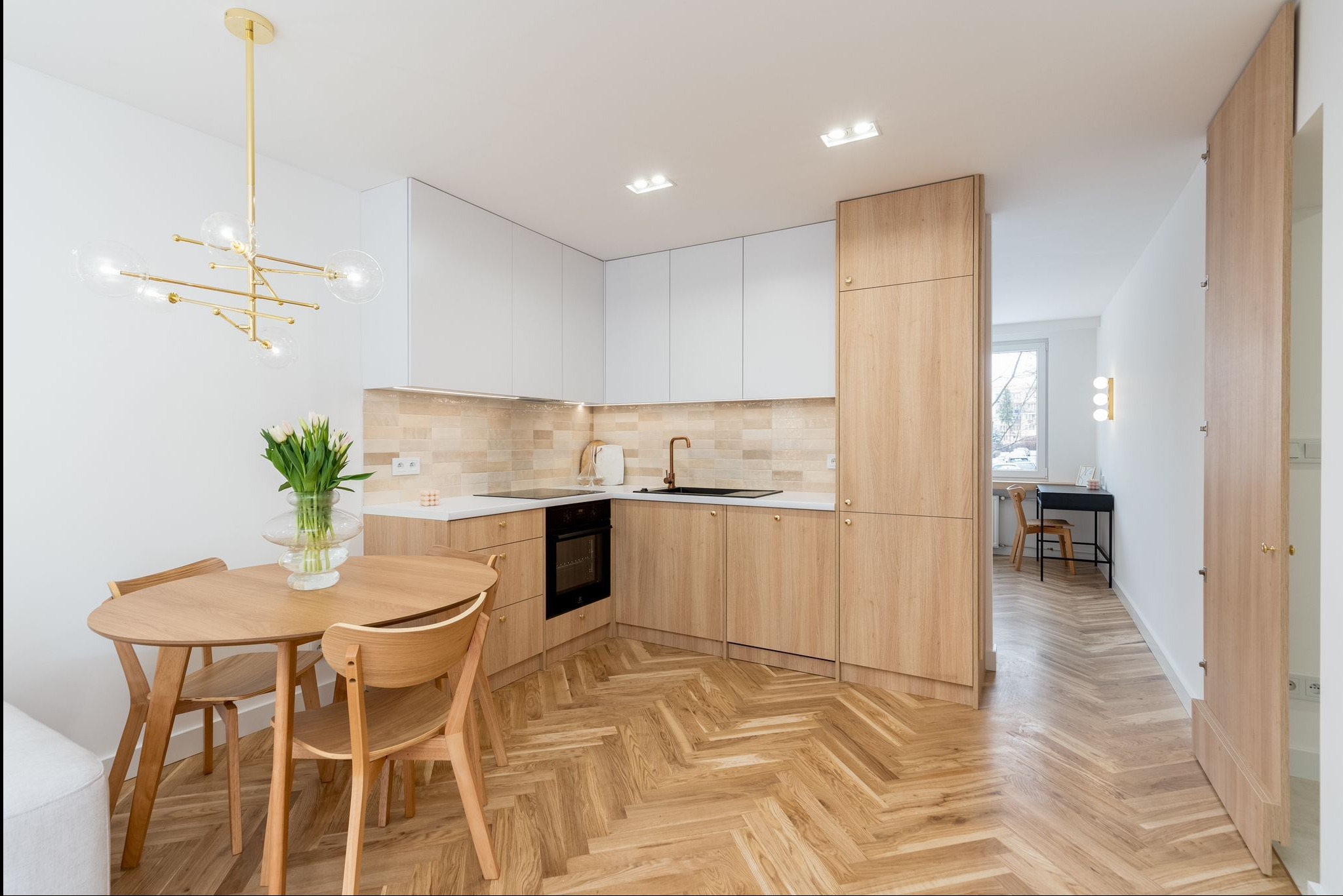 Jasna kuchnia z drewnianą podłogą ułożoną w jodełkę, białe szafki górne i drewniane dolne. Na stole wazon z tulipanami i złoty żyrandol.