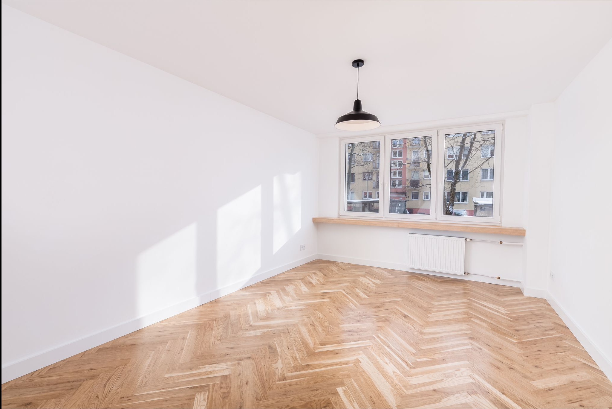 Jasne, puste wnętrze z parkietem w jodełkę, białymi ścianami i oknem z widokiem na budynek. Czarna lampa wisząca dodaje kontrastu. Minimalistyczny design.