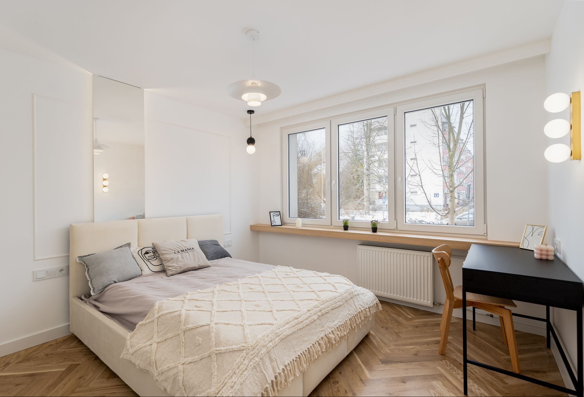 Jasna sypialnia z łóżkiem, biurkiem i dużym oknem. Parkiet w jodełkę, minimalistyczne lampy i lustro tworzą przytulną, nowoczesną przestrzeń.