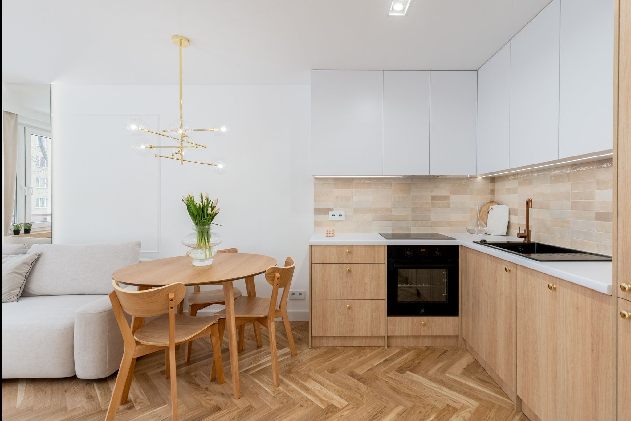 Jasne wnętrze: drewniany stół z tulipanami, minimalistyczna kuchnia z białymi szafkami i drewnianymi frontami, parkiet w jodełkę. Złoty żyrandol dodaje elegancji.