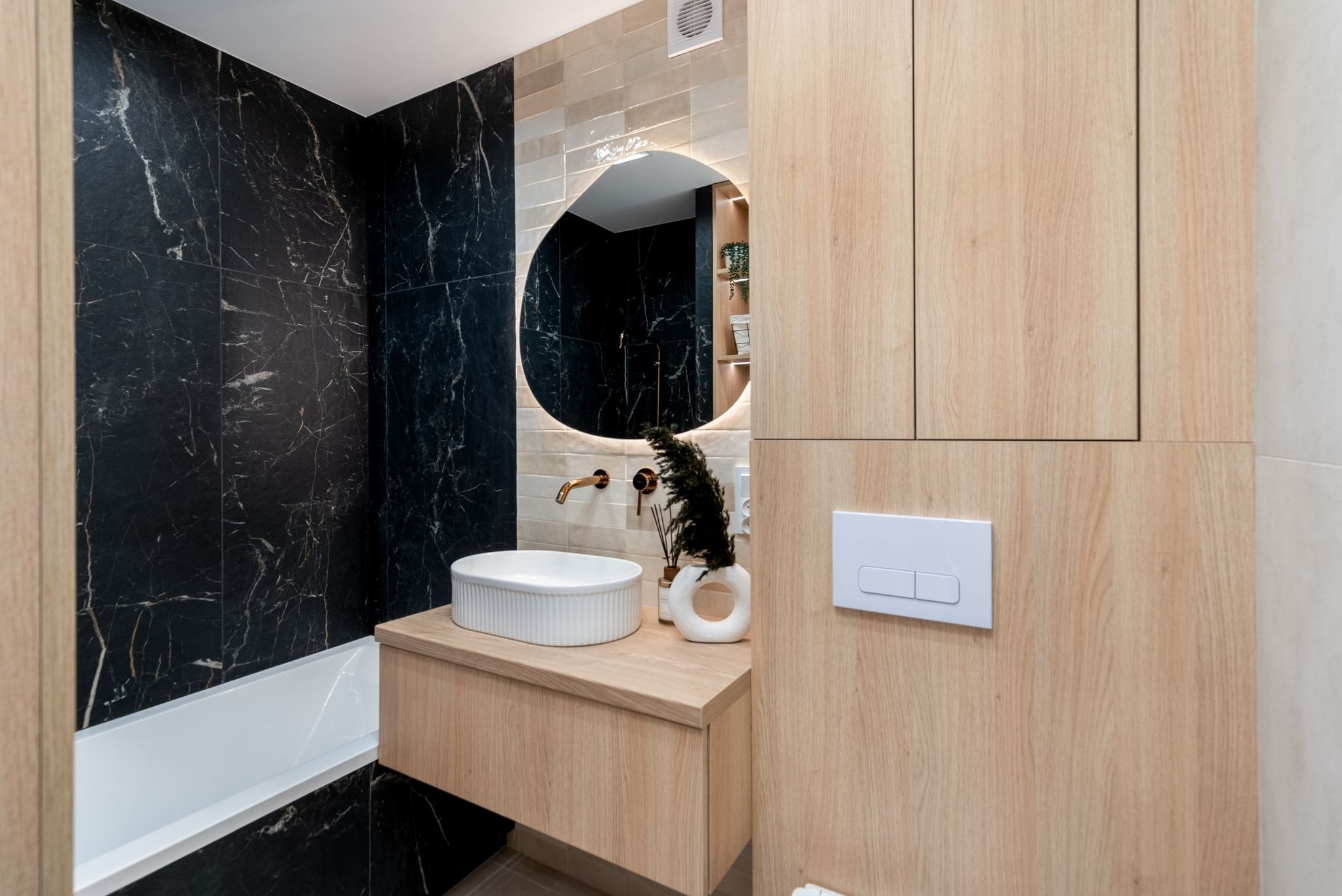 Nowoczesna łazienka z czarnymi marmurowymi płytkami, okrągłym lustrem z podświetleniem LED i drewnianą szafką pod umywalką.