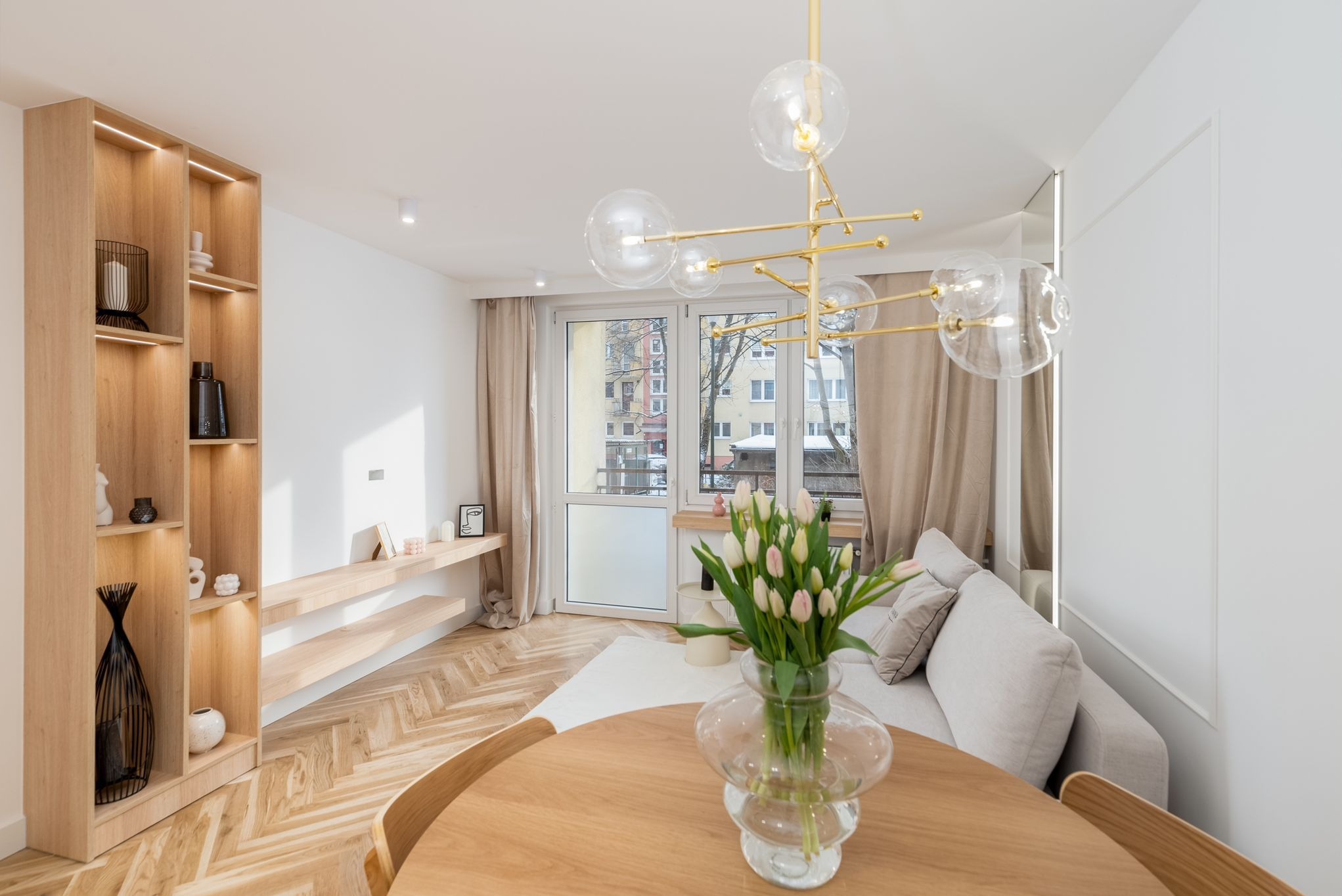 Jasne wnętrze salonu z drewnianą podłogą w jodełkę, regałem z oświetleniem LED i nowoczesną lampą wiszącą. Na stole wazon z tulipanami.
