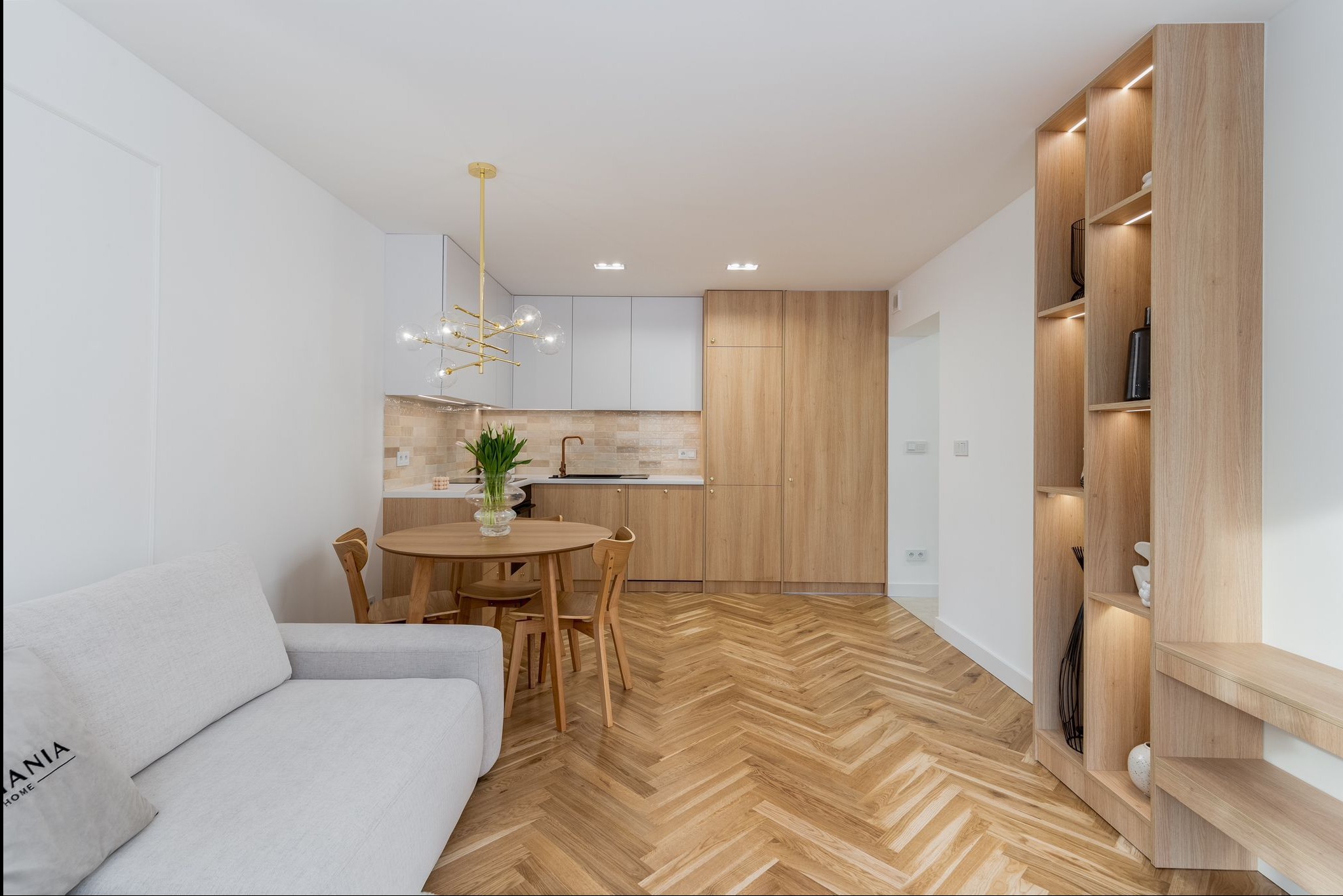 Jasne, nowoczesne wnętrze mieszkania z parkietem w jodełkę, okrągłym stołem, szarą kanapą i zabudową kuchenną z elementami drewna. Widok na regał z podświetleniem.