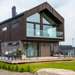 HAUKON Sp z.o.o firma budowlana - Nowoczesny dom z ciemnego drewna w Warszawie, z dużymi oknami i balkonem z przeszkloną balustradą, otoczony zielonym trawnikiem i młodymi drzewkami.