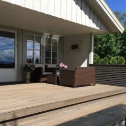 Zadaszony taras domu z drewnianą podłogą, meblami ogrodowymi z rattanu, jacuzzi po lewej stronie i dekoracyjną donicą z roślinami, widok na zieleń w tle.