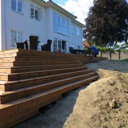 HAUKON Sp z.o.o firma budowlana - Drewniany taras schodkowy przed domem jednorodzinnym, mężczyzna w niebieskiej koszulce pracuje przy ziemi, widoczny stół i krzesła na tarasie.