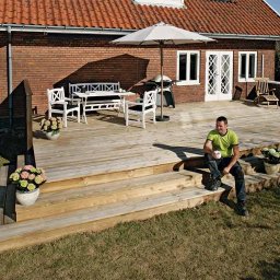 HAUKON Sp z.o.o firma budowlana - Ceglany dom z drewnianym tarasem, na którym siedzi mężczyzna w zielonej koszulce, obok białe meble ogrodowe i parasol.