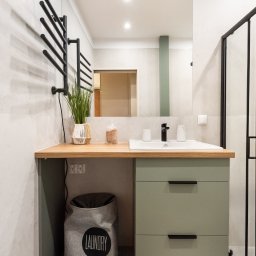 HAUKON Sp z.o.o firma budowlana - Nowoczesna łazienka z szafką umywalkową w odcieniach zieleni, drewnianym blatem, czarną armaturą i grzejnikiem drabinkowym; dekoracje obejmują roślinę w plecionym wazonie i pojemnik na sól...