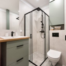 HAUKON Sp z.o.o firma budowlana - Nowoczesna łazienka w odcieniach szarości i zieleni z czarnymi akcentami, obejmująca kabinę prysznicową z czarną ramą, umywalkę z drewnianym blatem i szafką w kolorze oliwkowym oraz podwieszaną...