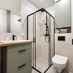 Nowoczesna łazienka w odcieniach szarości i zieleni z czarnymi akcentami, obejmująca kabinę prysznicową z czarną ramą, umywalkę z drewnianym blatem i szafką w kolorze oliwkowym oraz podwieszaną...