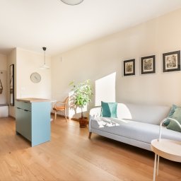 HAUKON Sp z.o.o firma budowlana - Jasne wnętrze mieszkania z drewnianą podłogą, turkusową wyspą kuchenną, białym narożnikiem z zielonymi poduszkami i roślinami doniczkowymi.