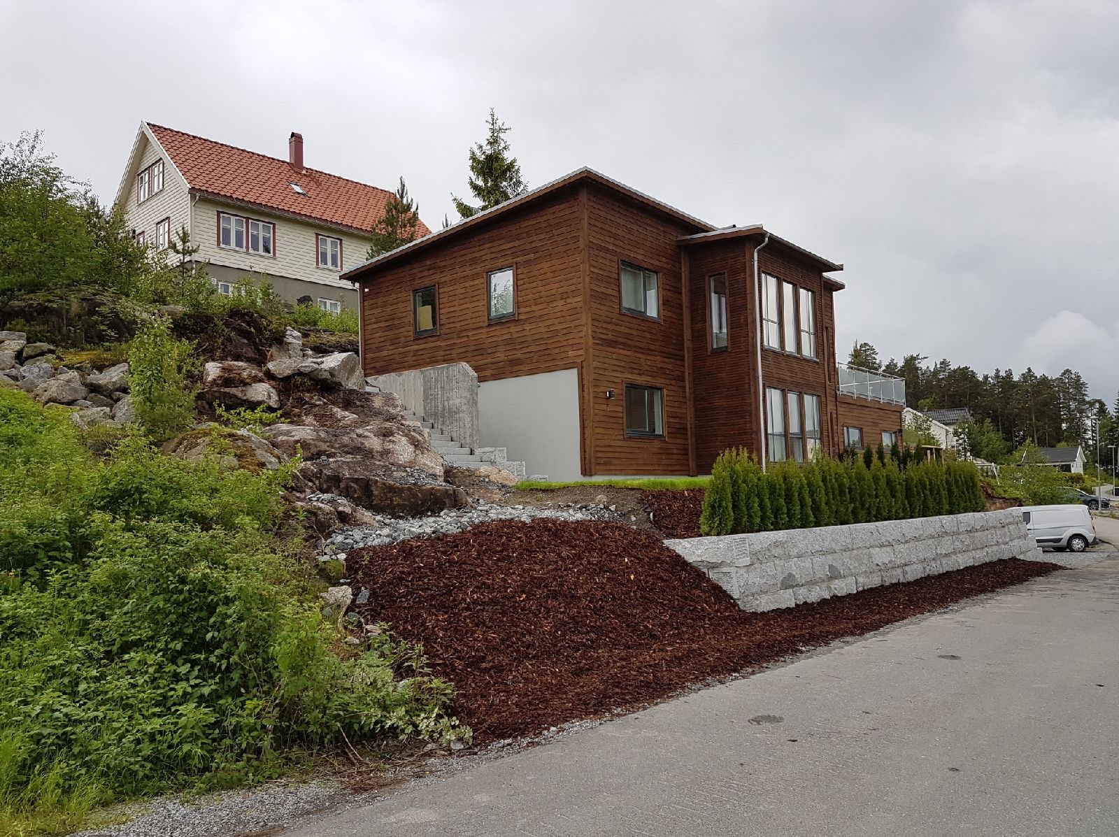 Dwukondygnacyjny dom z drewnianą elewacją, zbudowany na wzniesieniu z kamiennymi schodami i ozdobną korą drzewną, widoczny na tle pochmurnego nieba i sąsiedniego domu.