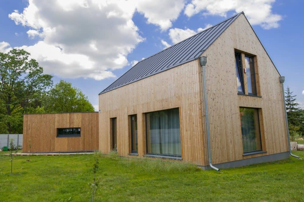 Nowoczesny dom szkieletowy z jasną, drewnianą elewacją pionową, grafitowym dachem i minimalistycznymi oknami na tle błękitnego nieba z chmurami.