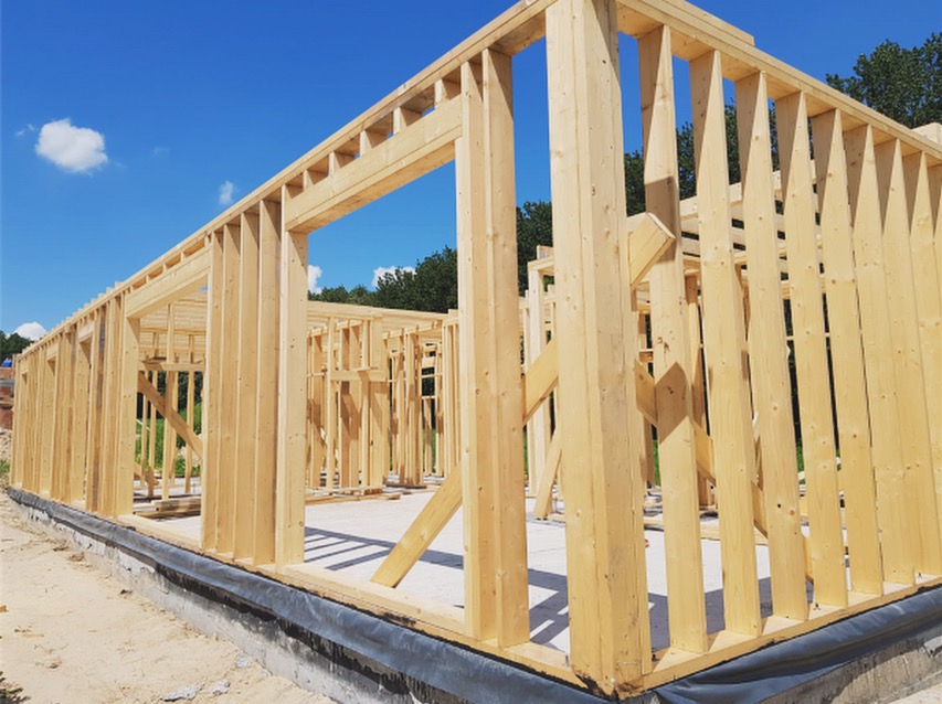 Surowy szkielet drewnianego domu w trakcie budowy, widoczne pionowe i poziome elementy konstrukcyjne, fundament z izolacją, błękitne niebo w tle.