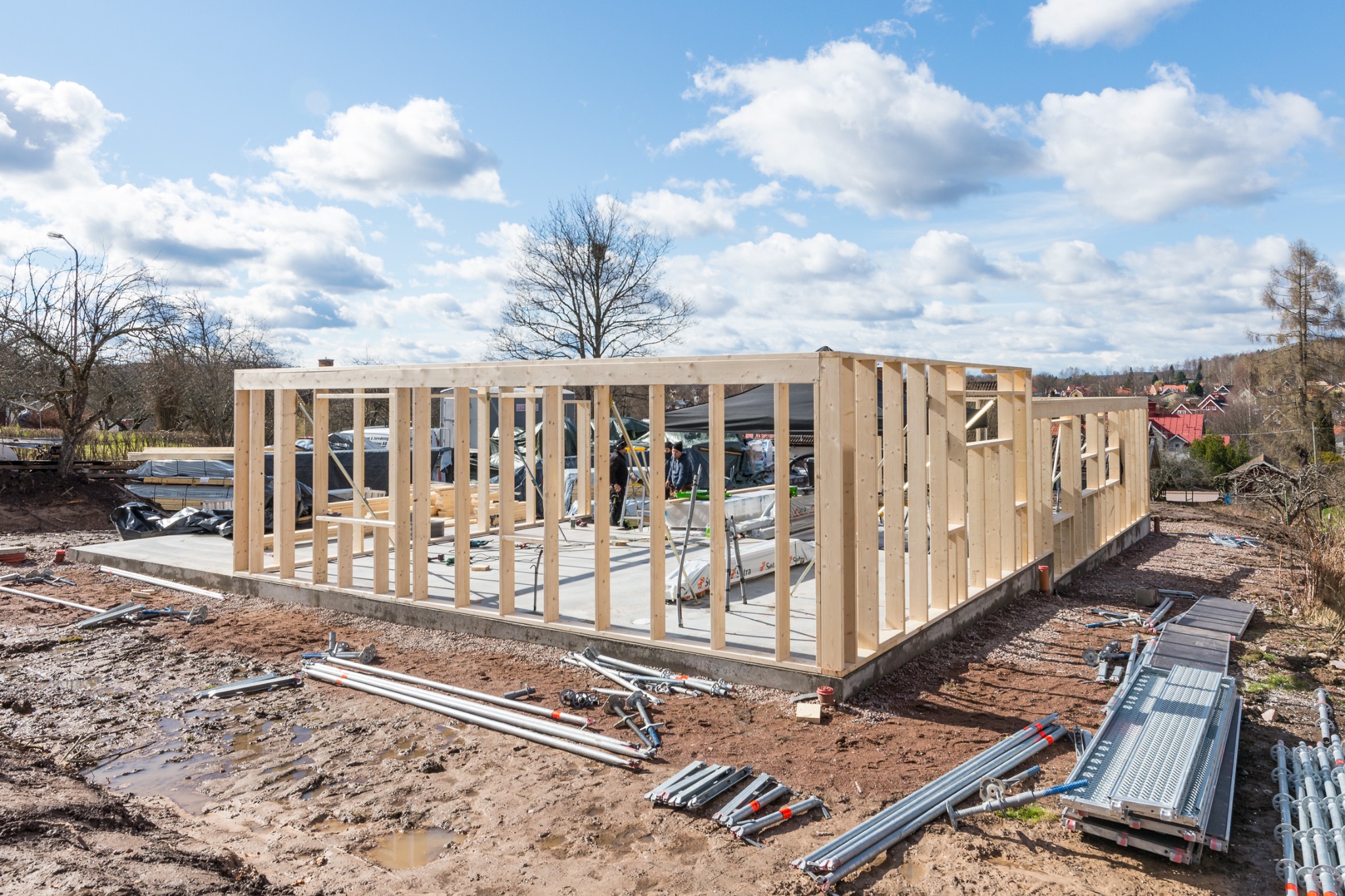 Wznoszenie drewnianej konstrukcji domu szkieletowego na fundamencie, widoczne rusztowania, podpory i narzędzia na błotnistym terenie, błękitne niebo z chmurami w tle.