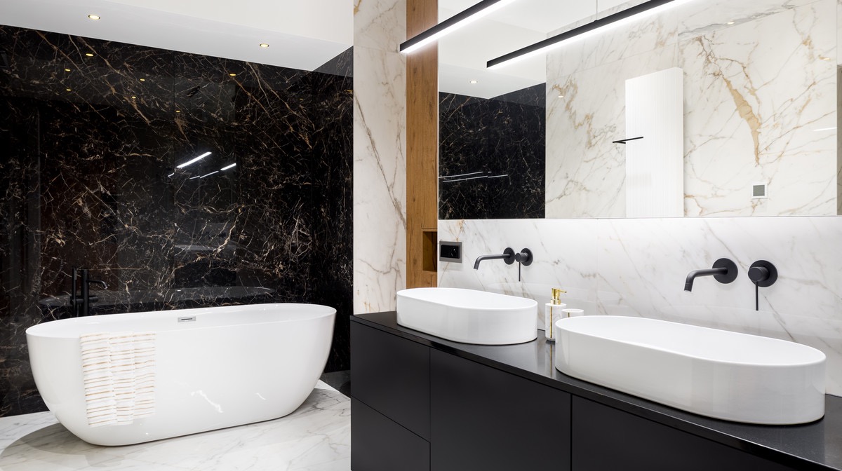 Elegancka łazienka z czarno-białymi marmurowymi płytkami, owalną białą wanną z ręcznikiem i dwiema owalnymi umywalkami nablatowymi z czarną armaturą.