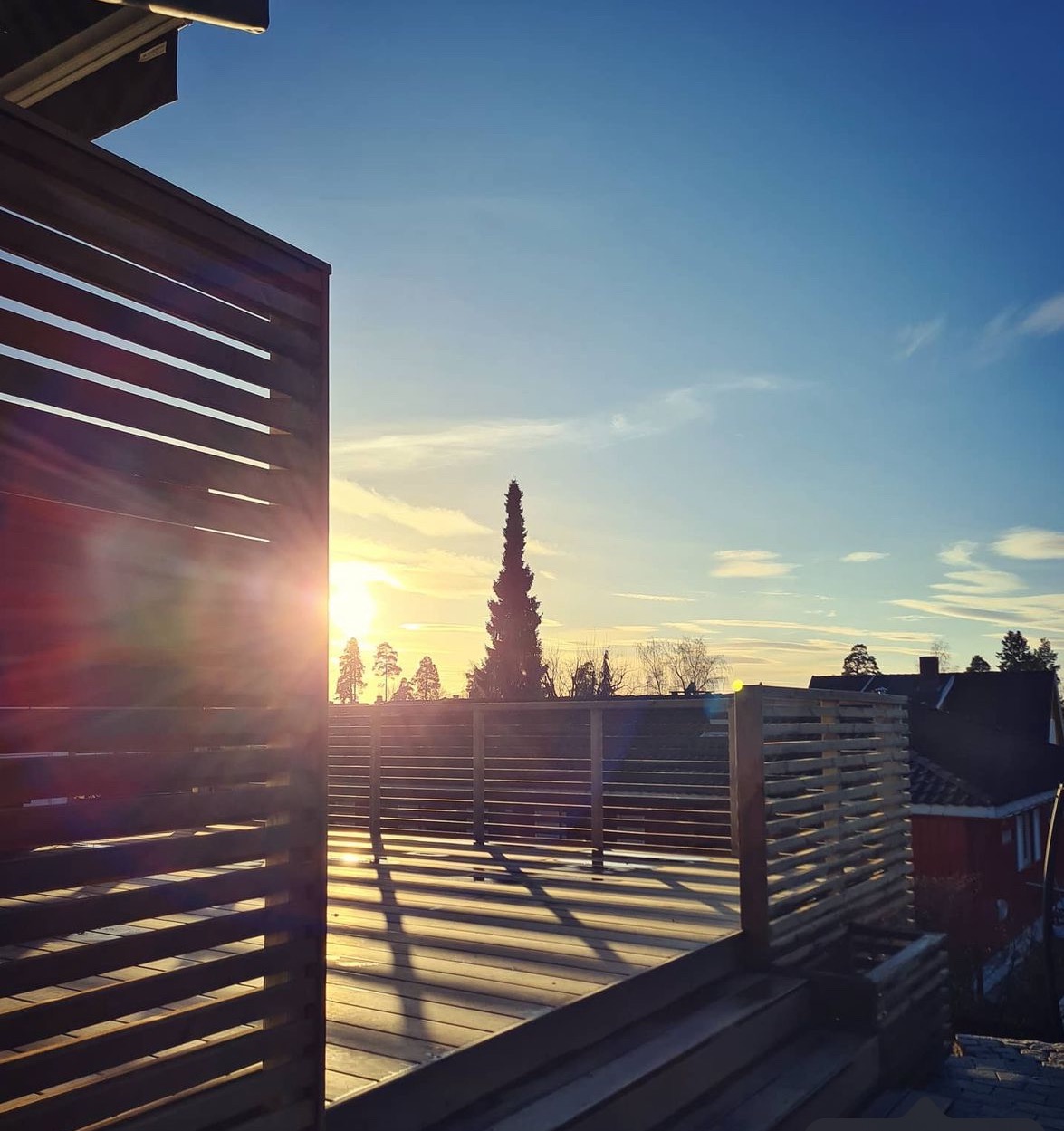 Drewniany taras z ażurową zabudową w promieniach zachodzącego słońca, widok na okoliczne drzewa i budynki.