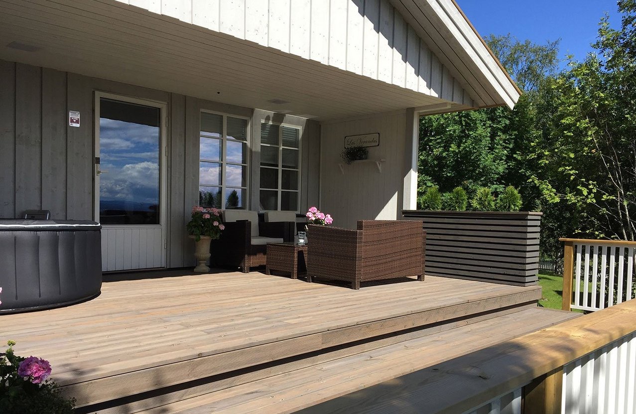 Zadaszony taras domu z drewnianą podłogą, meblami ogrodowymi z rattanu, jacuzzi po lewej stronie i dekoracyjną donicą z roślinami, widok na zieleń w tle.