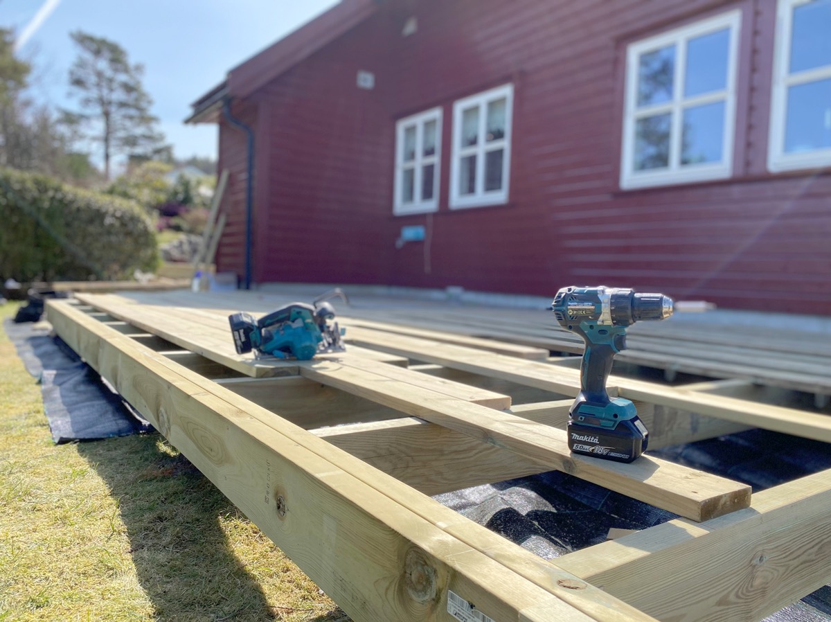 Budowa drewnianego tarasu przy czerwonym domu z białymi oknami; widoczne narzędzia: wiertarka i piła akumulatorowa.