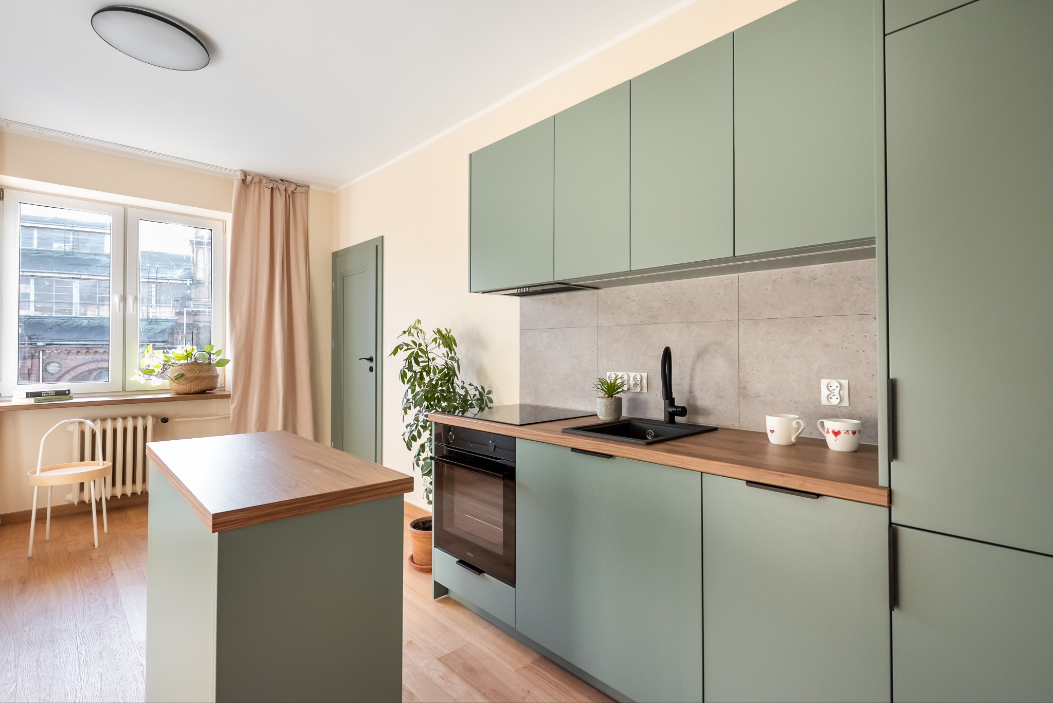 Jasna kuchnia w odcieniach zieleni z drewnianym blatem i wyspą, minimalistyczny design, roślina doniczkowa na blacie i przy oknie, dwa kubki z serduszkami, czarna armatura, okno z widokiem na budynek.