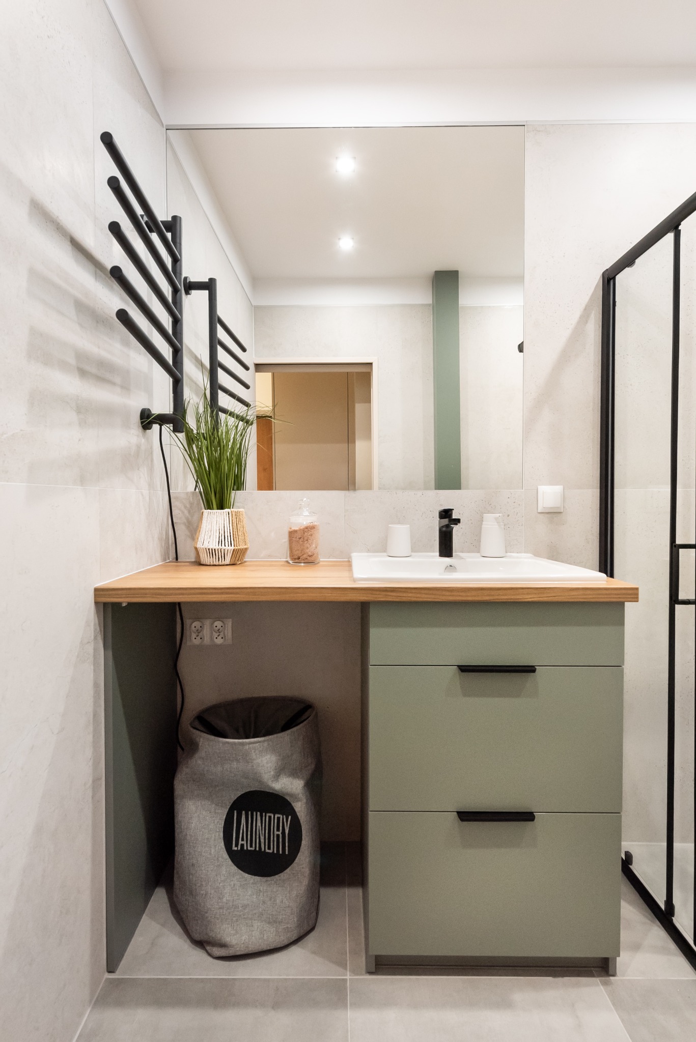 Nowoczesna łazienka z szafką umywalkową w odcieniach zieleni, drewnianym blatem, czarną armaturą i grzejnikiem drabinkowym; dekoracje obejmują roślinę w plecionym wazonie i pojemnik na sól...