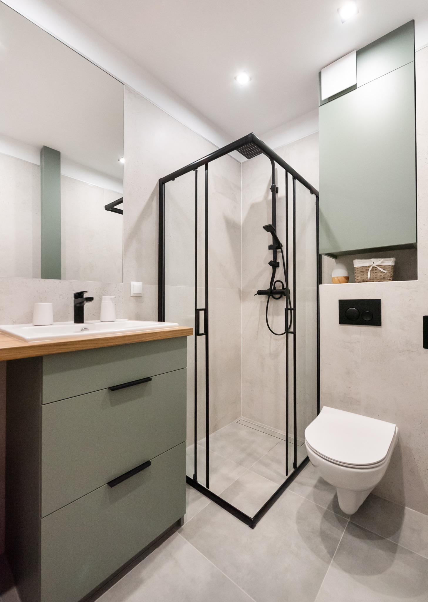 Nowoczesna łazienka w odcieniach szarości i zieleni z czarnymi akcentami, obejmująca kabinę prysznicową z czarną ramą, umywalkę z drewnianym blatem i szafką w kolorze oliwkowym oraz podwieszaną...